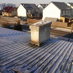 Blaszany dach pokryty szronem w porannym słońcu, z kominem w centrum kadru i widokiem na osiedle domów jednorodzinnych w tle.