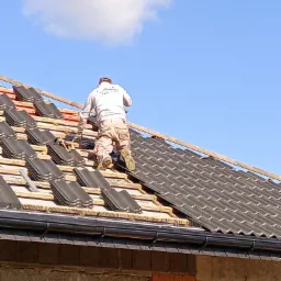 Pracownik w odzieży roboczej montuje ciemnoszarą dachówkę na drewnianej konstrukcji dachu budynku w trakcie budowy, widok od tyłu na tle błękitnego nieba.