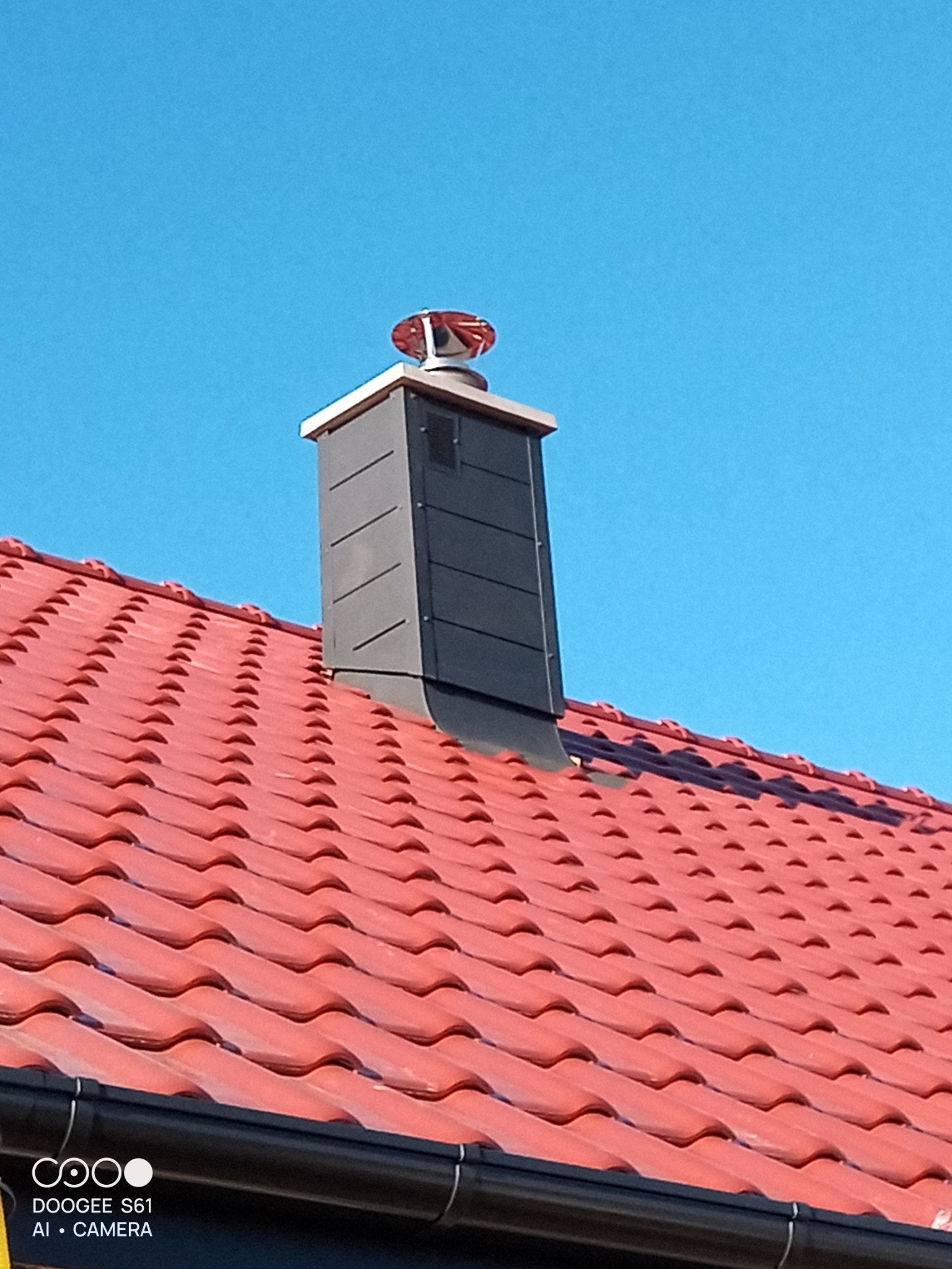 Czerwony dach z ceramicznej dachówki z czarnym kominem wentylacyjnym zwieńczonym wiatraczkiem na tle błękitnego nieba w słoneczny dzień.