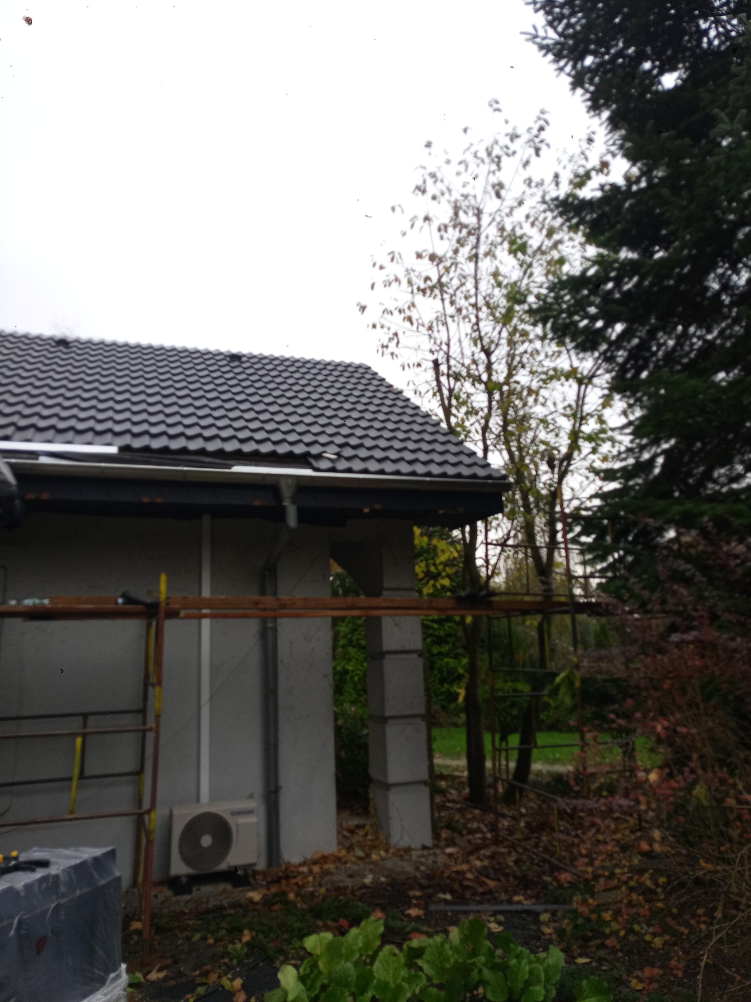 Szary dach z dachówki ceramicznej na budynku z szarą elewacją, widoczne rusztowanie i klimatyzator na zewnątrz. Jesienne otoczenie z drzewami i opadłymi liśćmi.