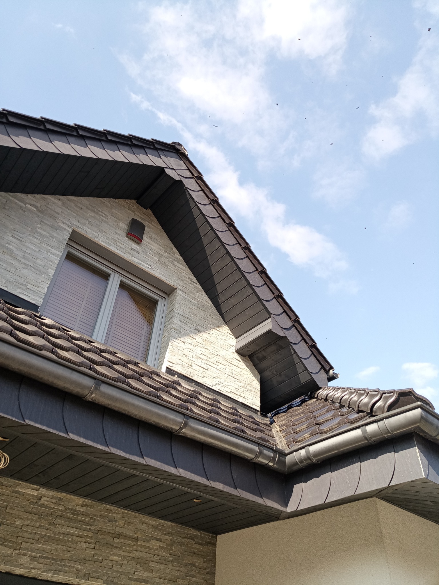 Nowoczesny dach z ciemnej dachówki ceramicznej i szarymi rynnami, widok z dołu na tle błękitnego nieba z chmurami. Elewacja budynku wykończona kamieniem.