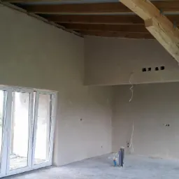 Wnętrze surowego domu po tynkowaniu maszynowym, widoczne gładkie, szare ściany, drewniane belki stropowe, okno balkonowe i instalacje hydrauliczne wystające z podłogi.