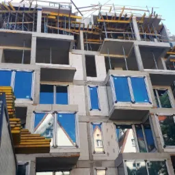 Wznoszenie wielopiętrowego budynku z betonu komórkowego, widoczne okna wstawione w surowy stan, szalunki stropowe na górnych kondygnacjach, kontener budowlany z boku.