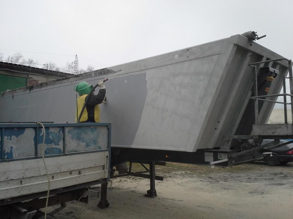 Pracownik w kasku i kamizelce ochronnej czyści strumieniowo przyczepę ciężarową, widoczna różnica w kolorze powierzchni przed i po zabiegu.