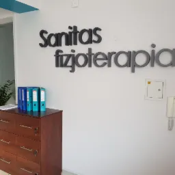 Recepcja gabinetu z napisem 'Sanitas fizjoterapia' na ścianie, brązowa komoda z segregatorami, roślina w białej donicy i biały domofon.