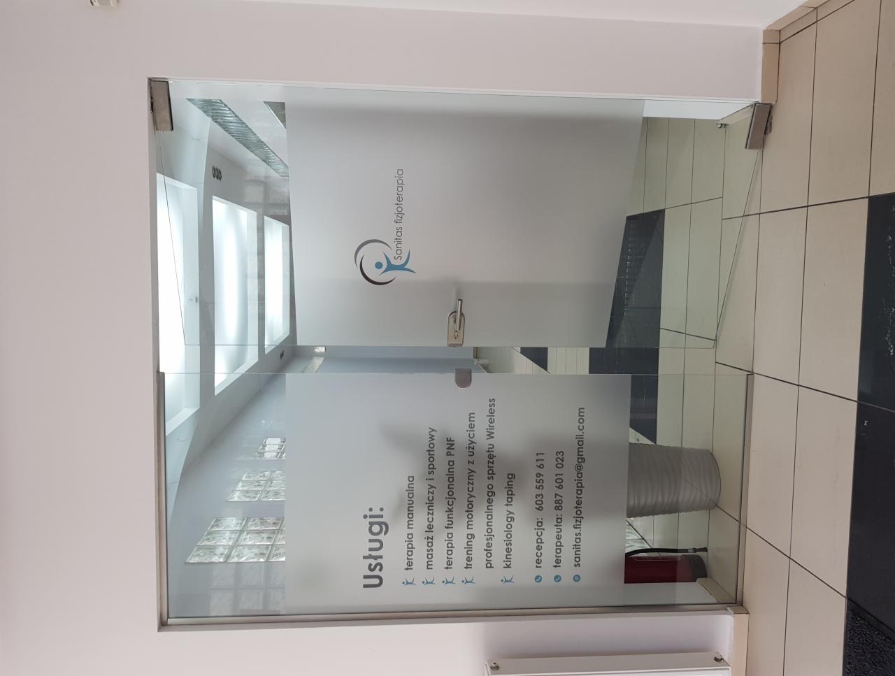 Szklane drzwi wejściowe do gabinetu fizjoterapii z naklejonym logo i listą oferowanych usług: terapia manualna, masaż leczniczy i sportowy, terapia funkcjonalna PNF, trening motoryczny z użyciem...