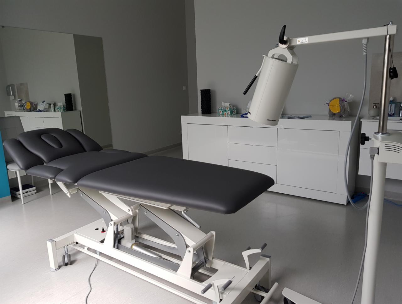 Profesjonalny stół rehabilitacyjny z regulowanym zagłówkiem i podnośnikiem elektrycznym, ustawiony w jasnym gabinecie z białymi meblami i szarymi ścianami.