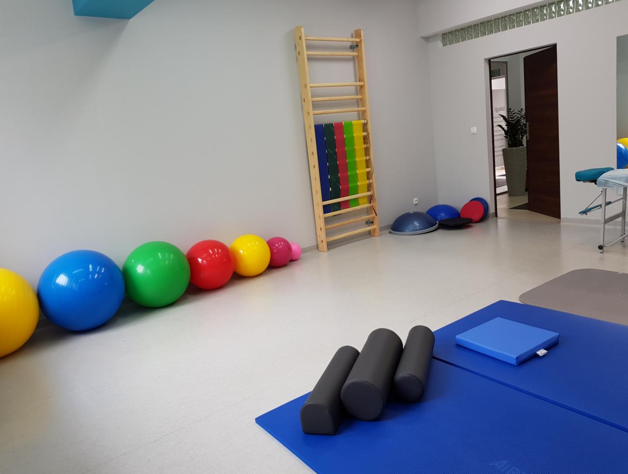 Jasne wnętrze gabinetu z drabinką gimnastyczną, kolorowymi piłkami rehabilitacyjnymi ułożonymi w rzędzie, wałkami do masażu, matami do ćwiczeń i stołem do masażu w tle.