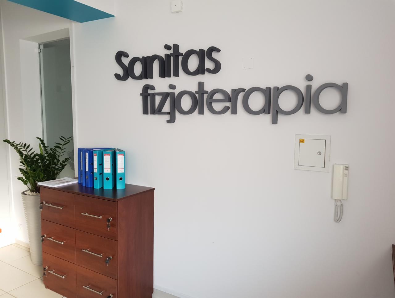 Recepcja gabinetu z napisem 'Sanitas fizjoterapia' na ścianie, brązowa komoda z segregatorami, roślina w białej donicy i biały domofon.
