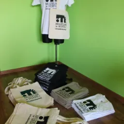 Prezentacja firmowych gadżetów Muzeum w Mokrej: biała koszulka na manekinie, beżowa torba bawełniana z nadrukiem logo, sterty koszulek w kolorach czarnym, beżowym i białym oraz złożone torby...