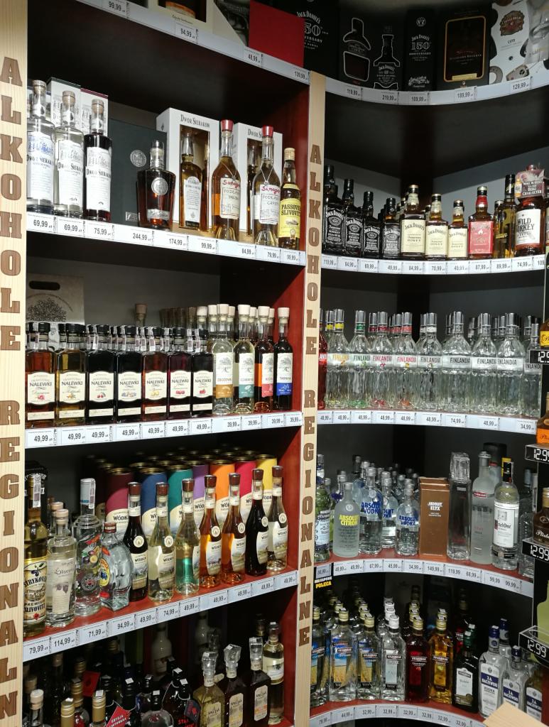 Półki w sklepie alkoholowym wypełnione różnorodnymi butelkami wódek, likierów i whisky, z cenami widocznymi na etykietach, oświetlone światłem sklepowym.