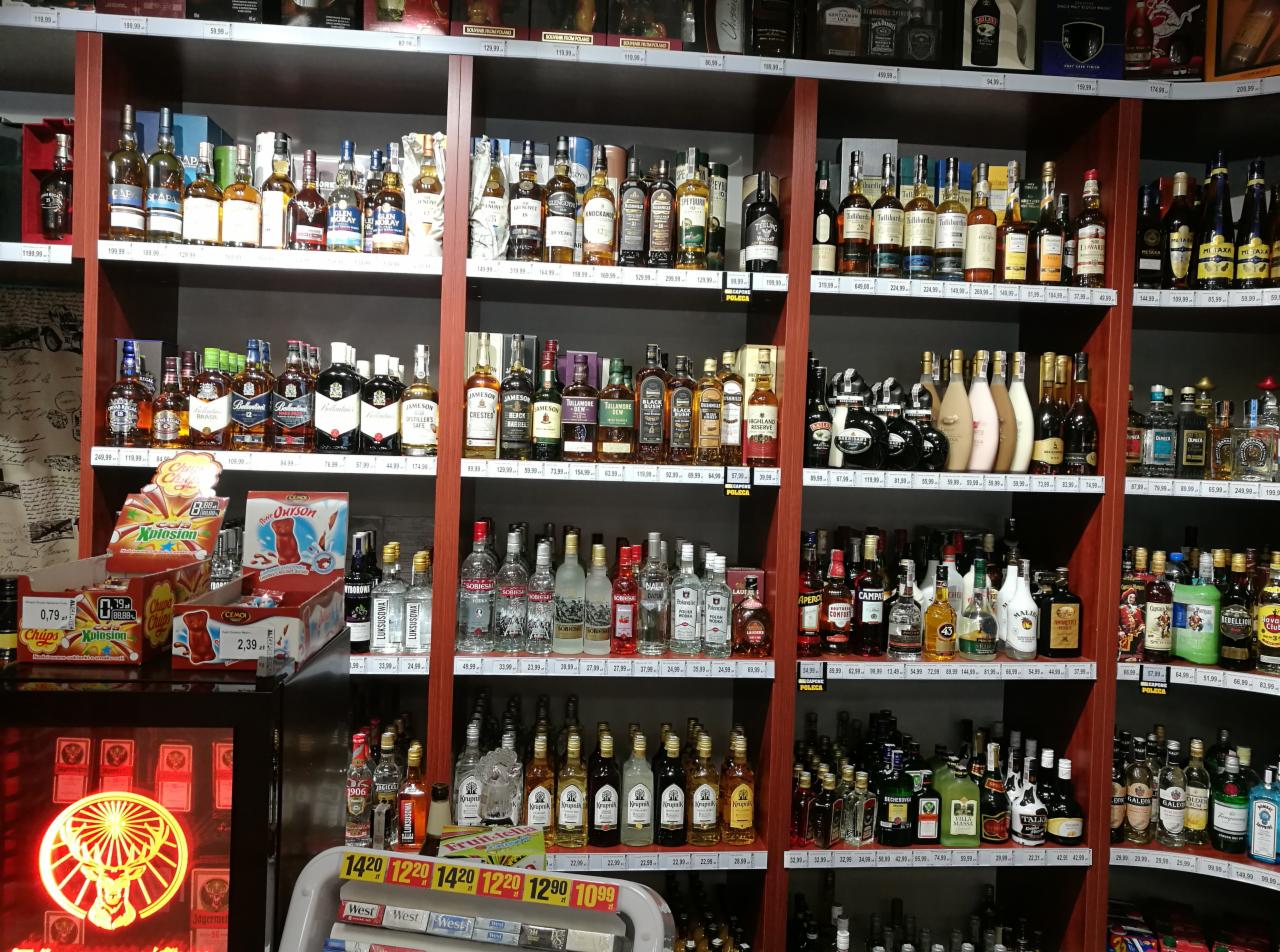 Półki sklepowe wypełnione różnorodnymi butelkami alkoholi, słodyczami i papierosami, z podświetlonym logo Jägermeister w lewym dolnym rogu.