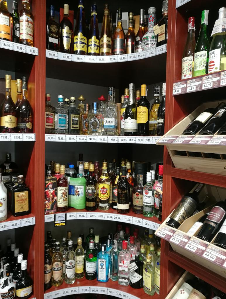Półki w sklepie z alkoholem, wypełnione różnorodnymi butelkami wódek, likierów i win, z widocznymi cenami na etykietach.