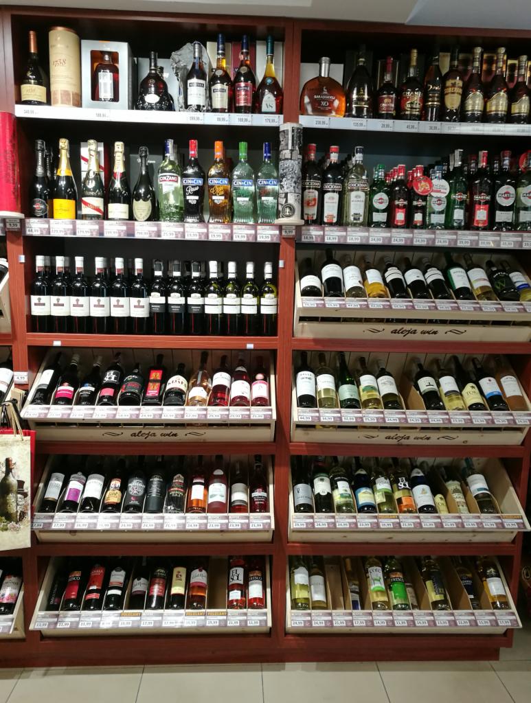 Półki sklepowe wypełnione różnorodnymi butelkami alkoholi, w tym win, likierów i wódek, z cenami widocznymi pod produktami.