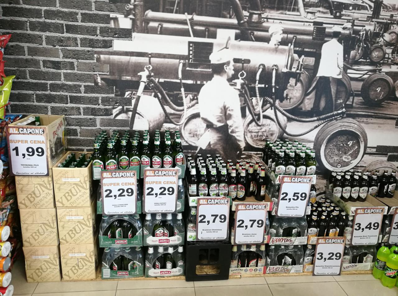 Ekspozycja różnych marek piwa (Perła, Namysłów, Braniewo) z cenami w sklepie, na tle ściany z motywem browaru.
