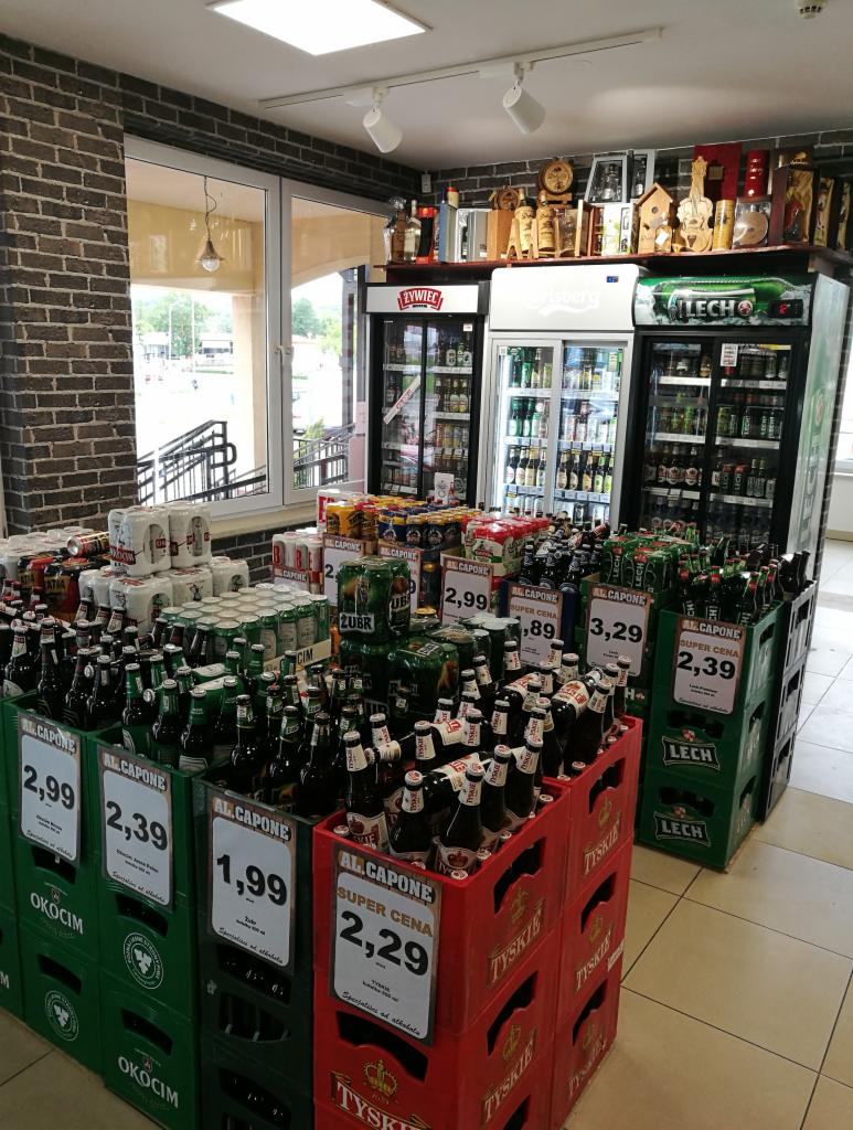 Wnętrze sklepu z napojami alkoholowymi, prezentujące ekspozycję piw w butelkach i puszkach, ułożonych w skrzynkach marek Okocim i Tyskie, z widocznymi cenami i lodówkami chłodniczymi w tle,...