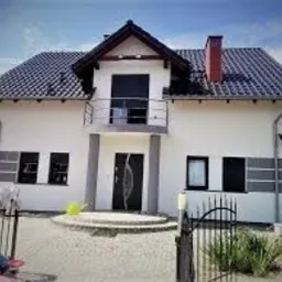 Dwukondygnacyjny dom z białą elewacją, ciemnym dachem i balkonem z czarną balustradą, widoczny z poziomu brukowanego podjazdu z czarną bramą wjazdową, w tle błękitne niebo.