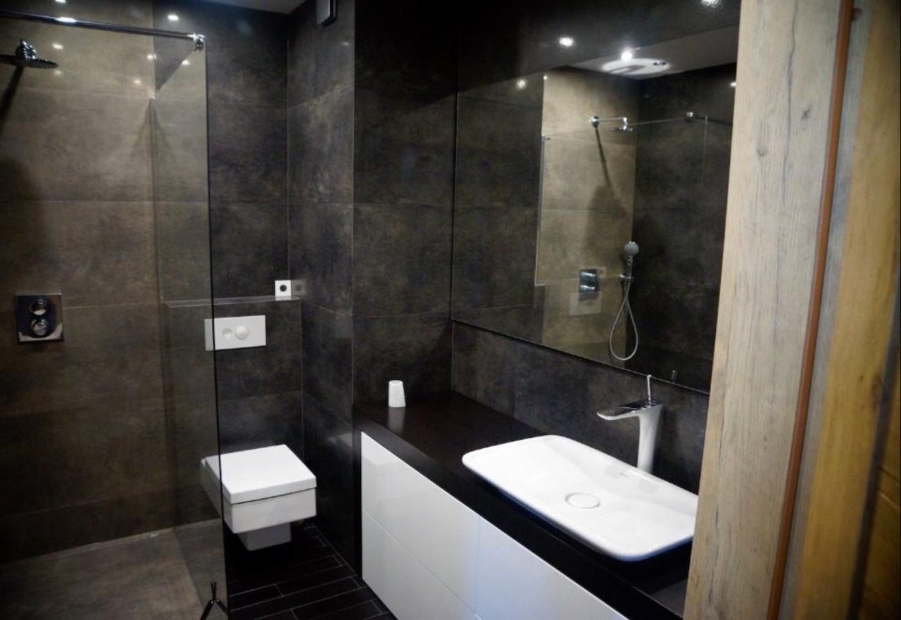 Nowoczesna łazienka z ciemnymi płytkami, kabiną prysznicową ze szklaną ścianką, podwieszaną toaletą i umywalką nablatową na białej szafce z ciemnym blatem.