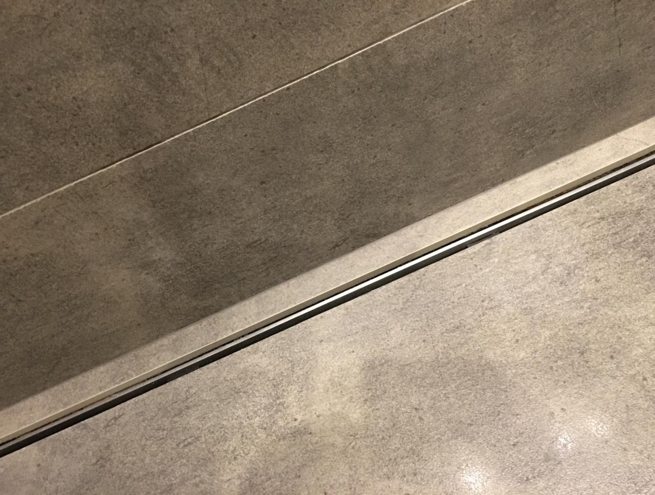 Szczegółowy widok na podłogę i ścianę wyłożone szarymi płytkami imitującymi beton, z widocznym odpływem liniowym z metalową listwą.