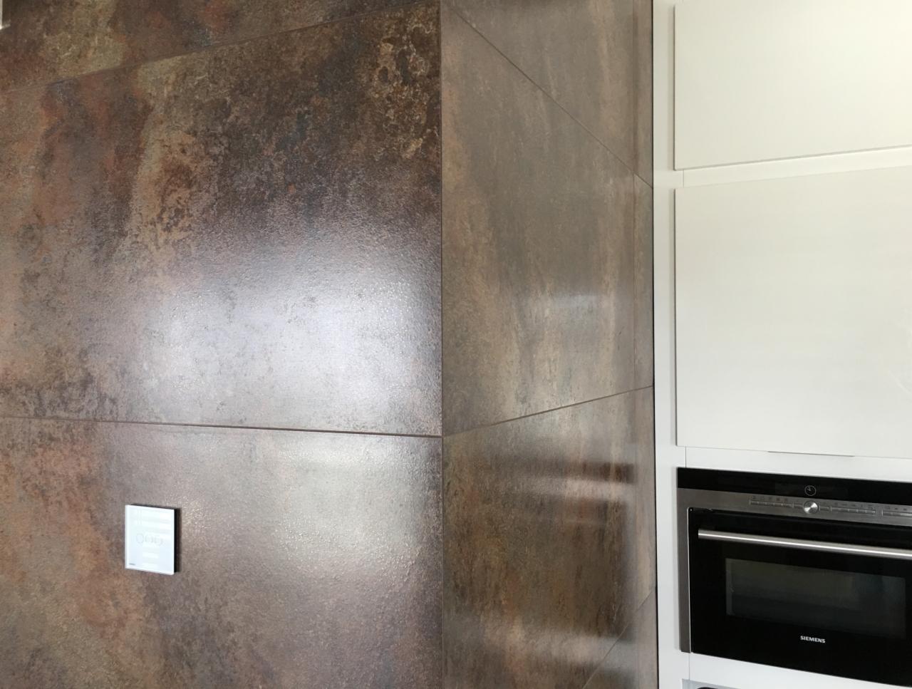 Fragment ściany wyłożonej brązowymi, błyszczącymi płytkami imitującymi rdzę, sąsiadujący z białymi szafkami kuchennymi i wbudowanym piekarnikiem marki Siemens.