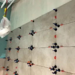 Ściana z jasnych płytek ceramicznych w trakcie układania, z użyciem systemu poziomowania płytek (klipsy i kliny), widoczny poziomica i worki z materiałem budowlanym w tle.