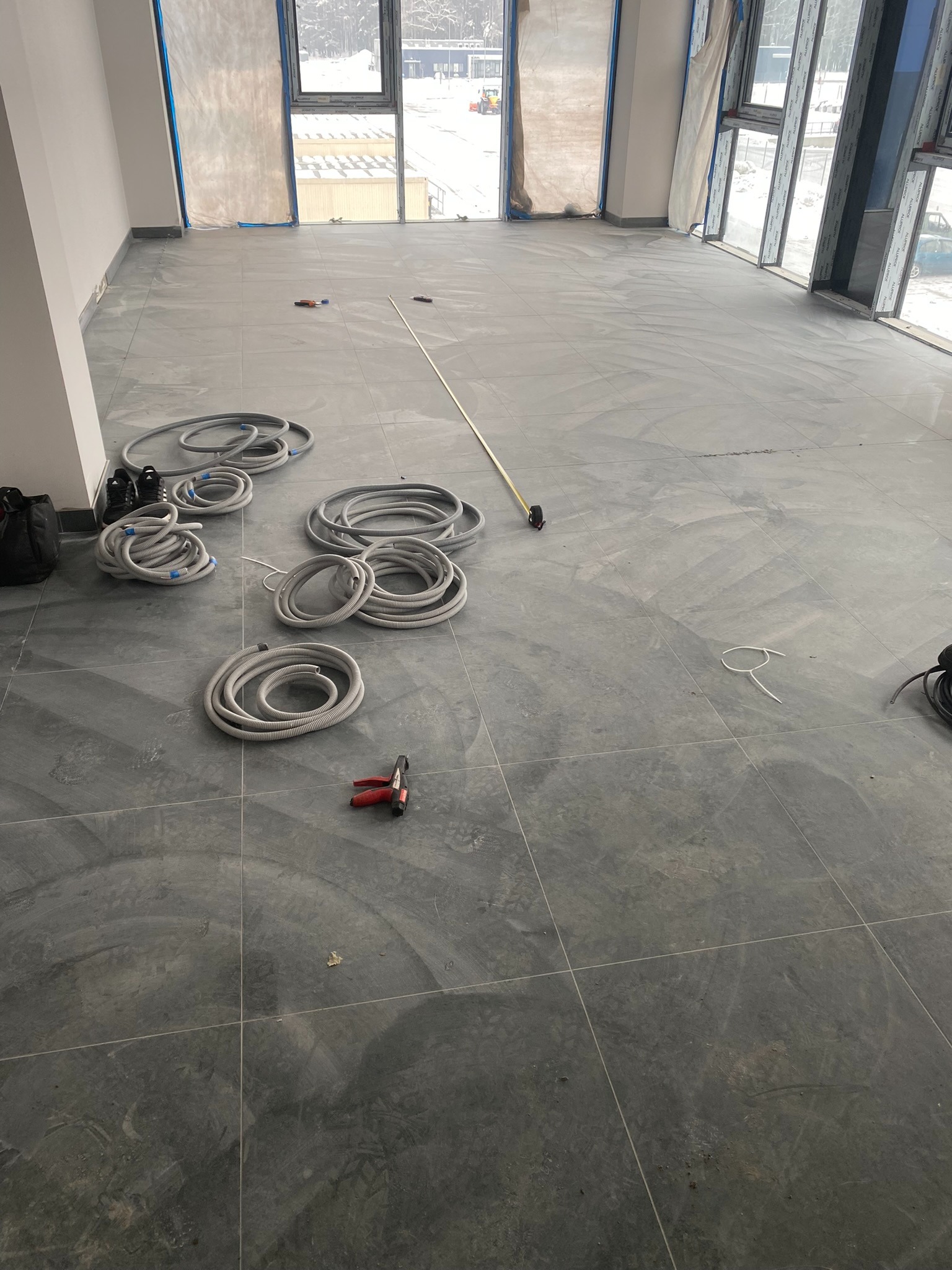 Szare płytki podłogowe w dużym, jasnym pomieszczeniu w trakcie prac wykończeniowych, widoczne zwinięte rury instalacyjne, narzędzia oraz okna zabezpieczone folią malarską.
