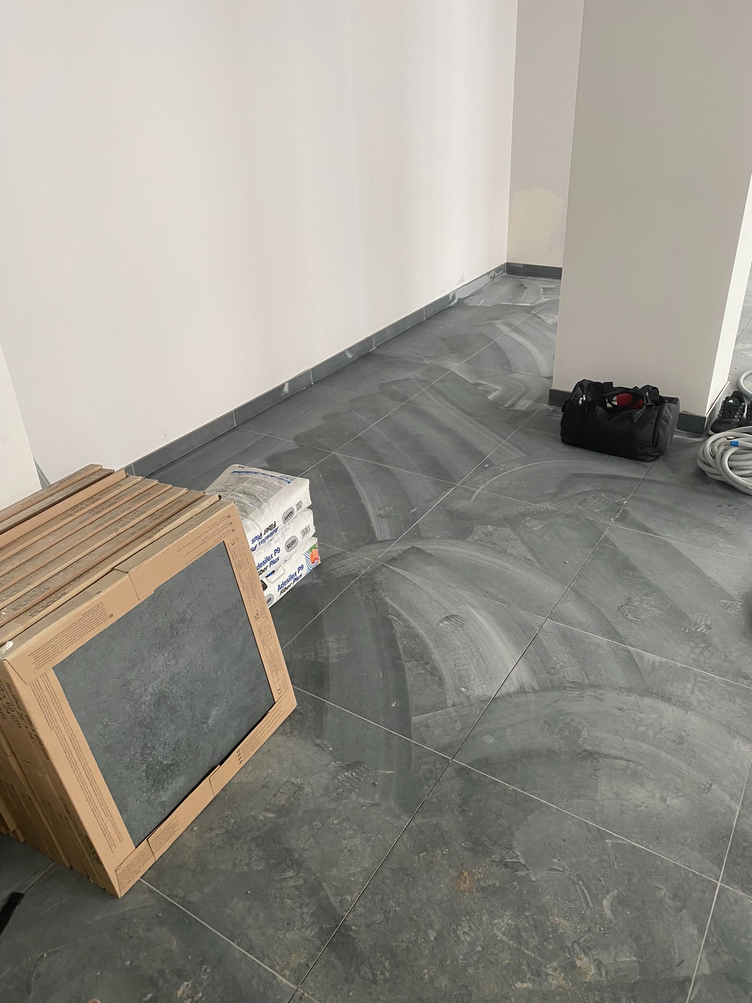 Szare płytki podłogowe po fugowaniu, z widocznymi śladami fugi, kartony z nowymi płytkami, torba z narzędziami i białe kable w rogu pomieszczenia.