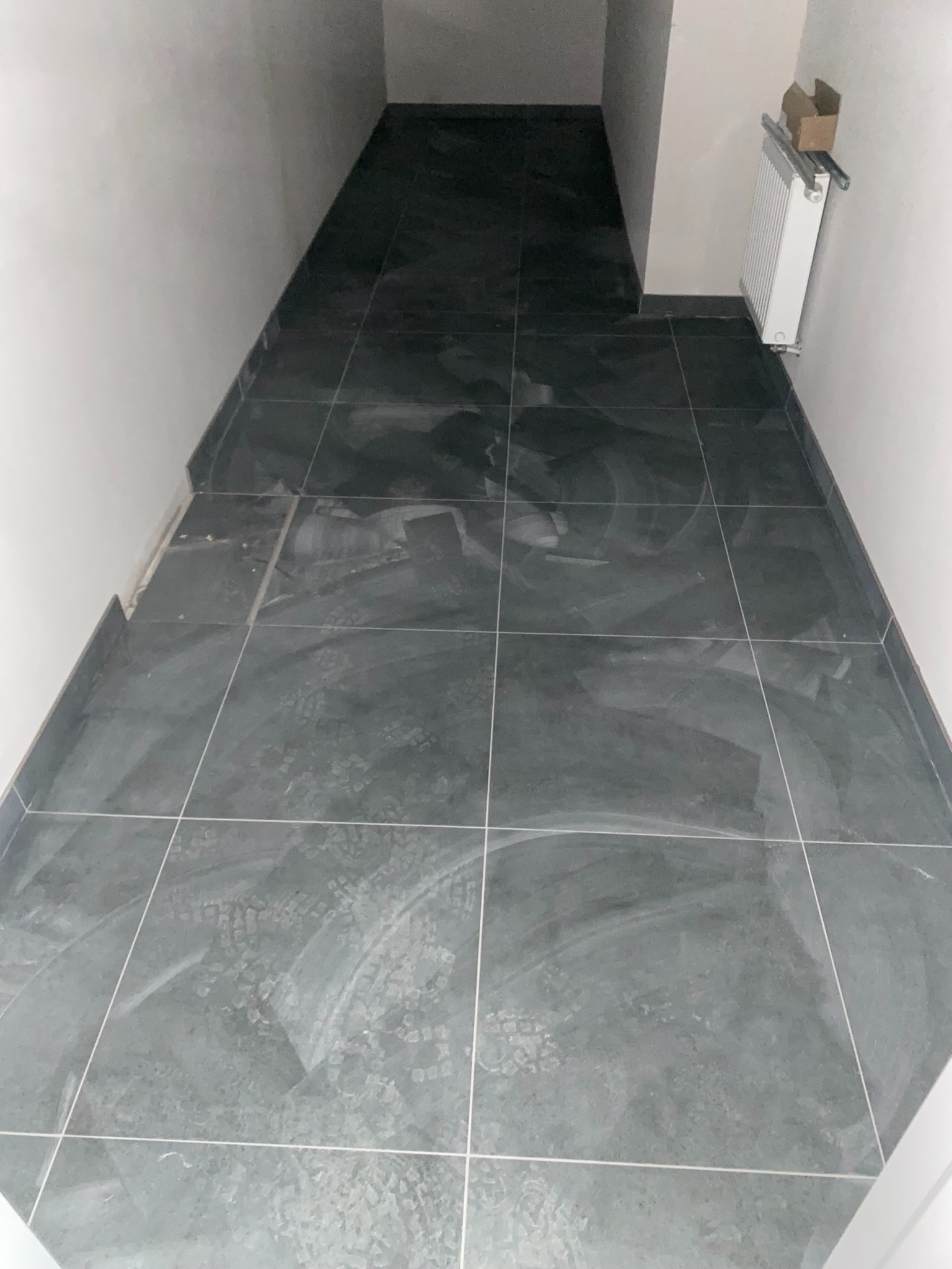 Ułożone ciemnoszare płytki podłogowe w wąskim korytarzu, widoczne ślady po butach, białe ściany, grzejnik z kartonem na górze.