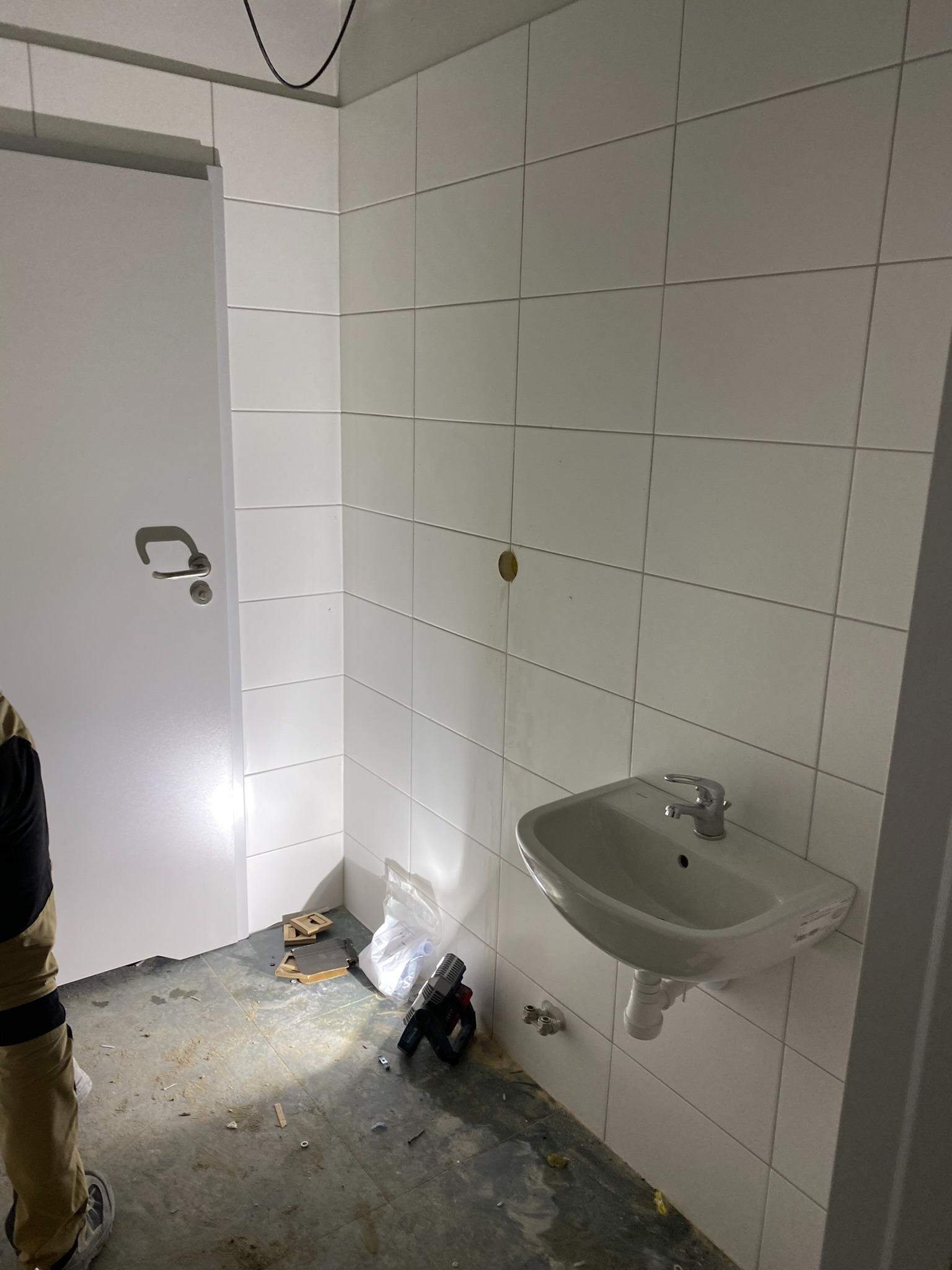 Wnętrze łazienki w trakcie prac: białe płytki na ścianach, zamontowana umywalka, widoczne narzędzia i materiały budowlane na podłodze, białe drzwi po lewej stronie.