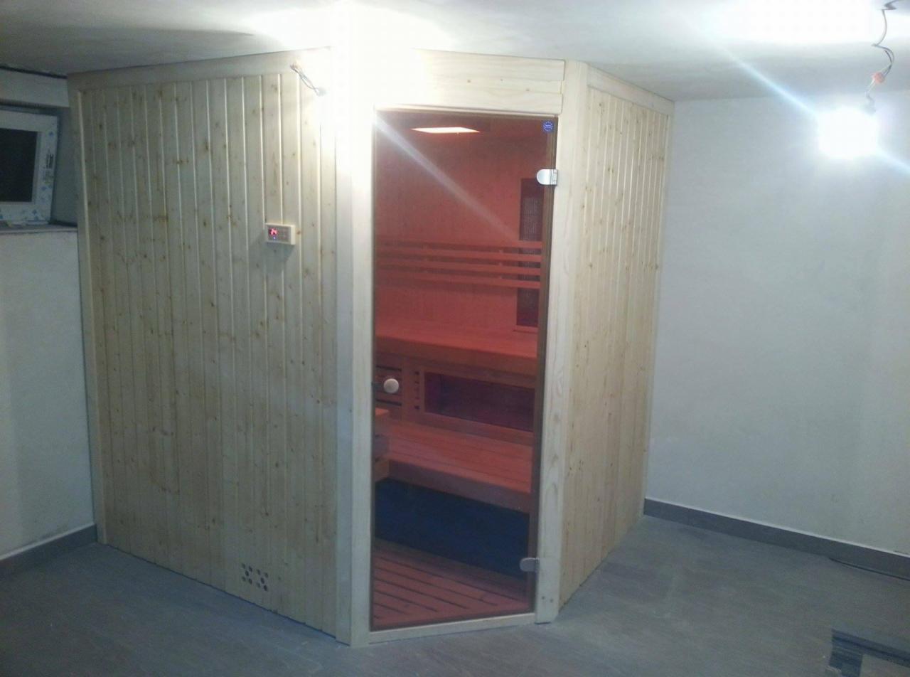 Kątowa kabina z jasnego drewna z przeszklonymi drzwiami, widoczne wewnętrzne ławy i panel sterowania temperaturą, surowy wystrój pomieszczenia.