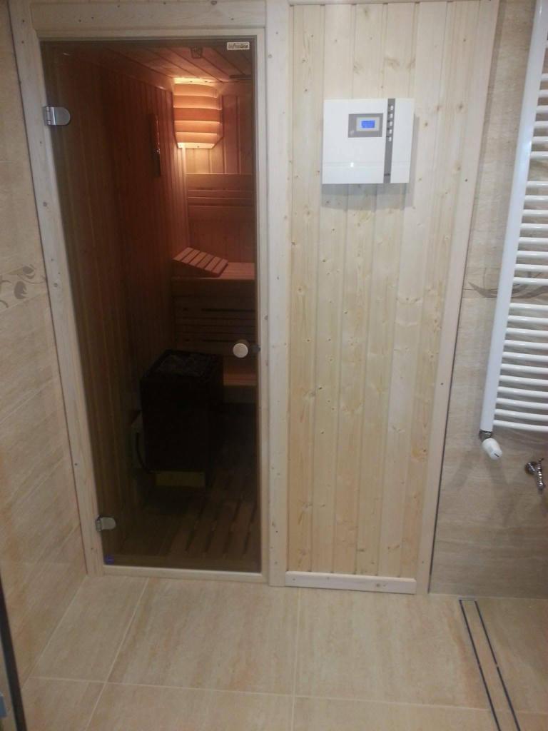 Wnętrze małej, domowej kabiny z przeszklonymi drzwiami, drewniane ławy i piec, na zewnątrz panel sterowania i grzejnik łazienkowy.