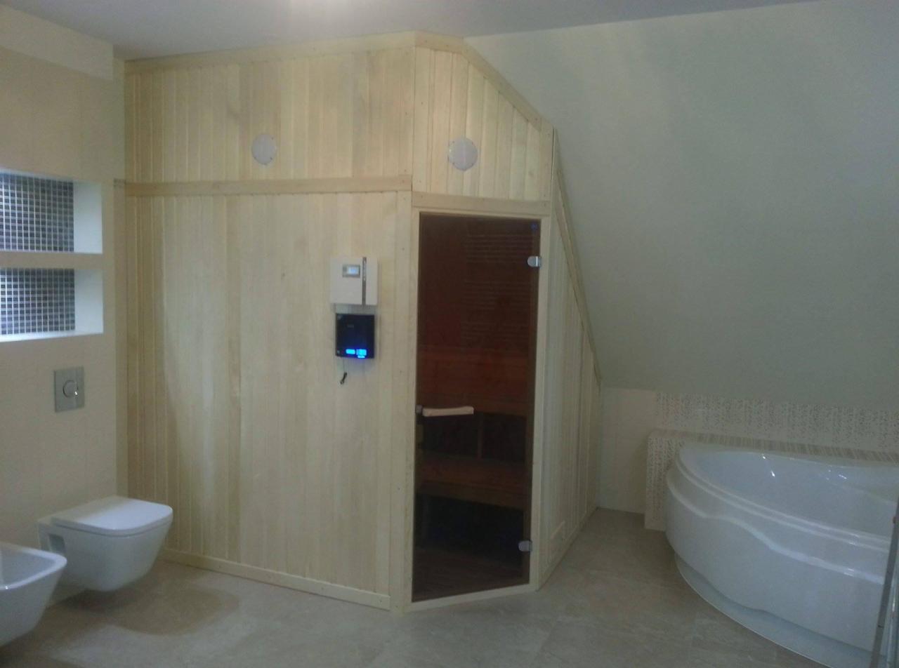 Jasna drewniana kabina z przeszklonymi drzwiami w pomieszczeniu z wanną i toaletą; panel sterowania na zewnątrz.