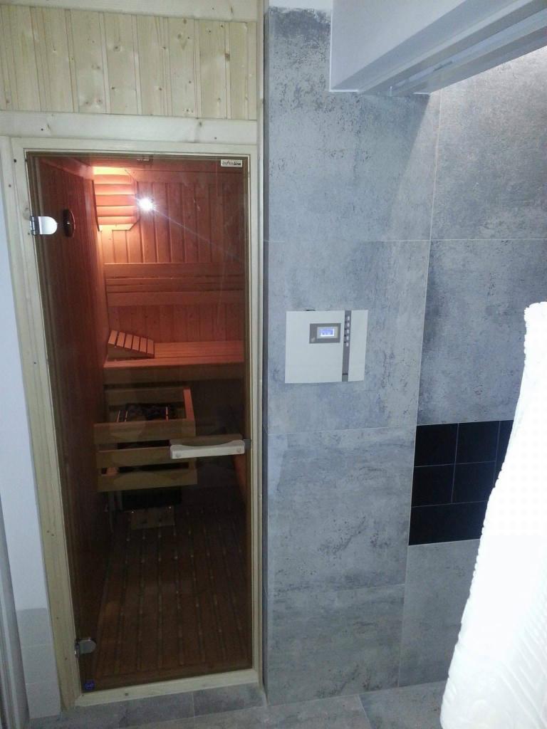 Wnętrze sauny z drewnianymi ławkami, widoczne przez szklane drzwi, zintegrowane z łazienką wykończoną szarymi płytkami imitującymi kamień i czarnymi akcentami.