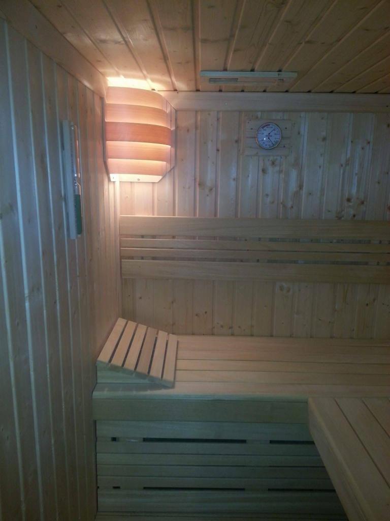 Wnętrze drewnianej kabiny z ławkami, oświetleniem ściennym i zegarem na ścianie.