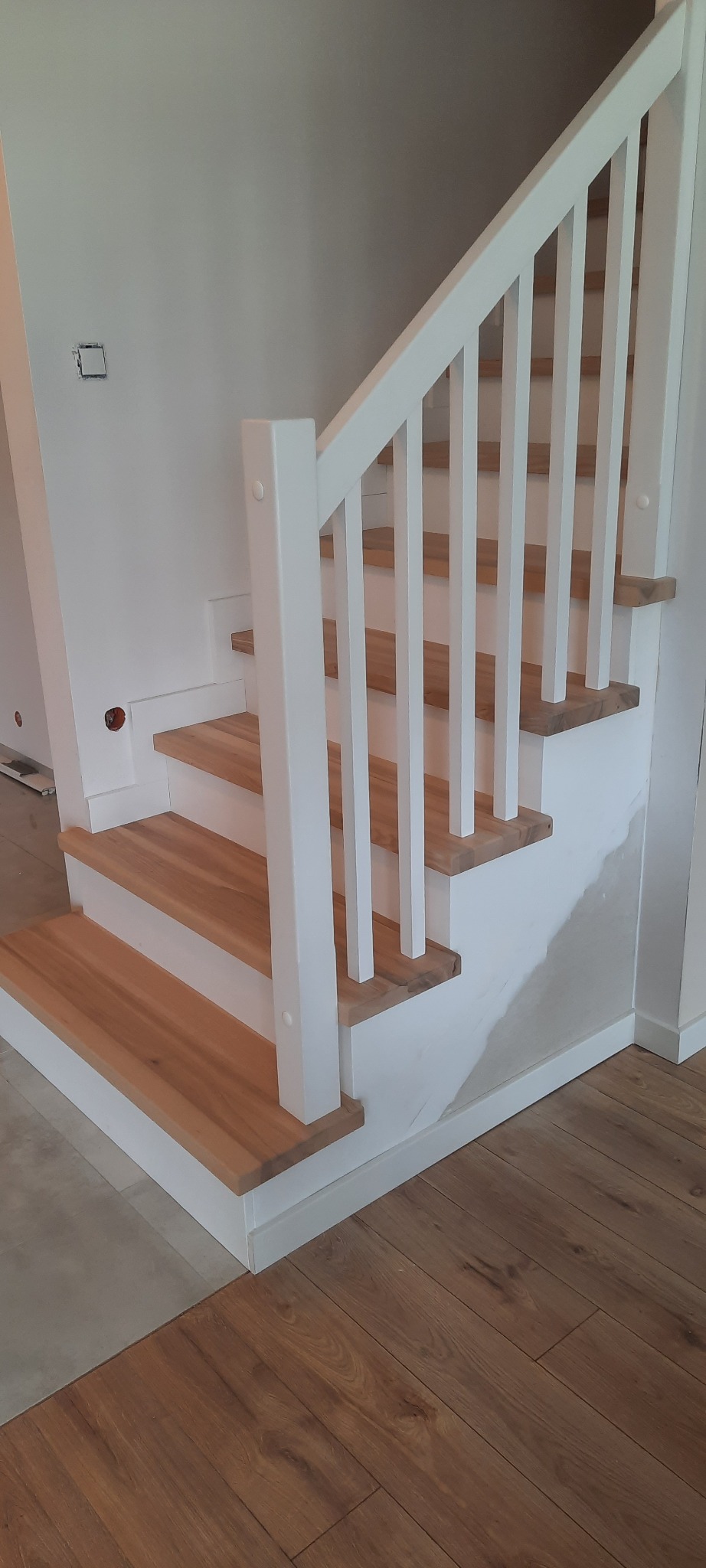 Jasne drewniane stopnie schodów z białymi podstopnicami i balustradą, ściana częściowo wykończona gładzią.
