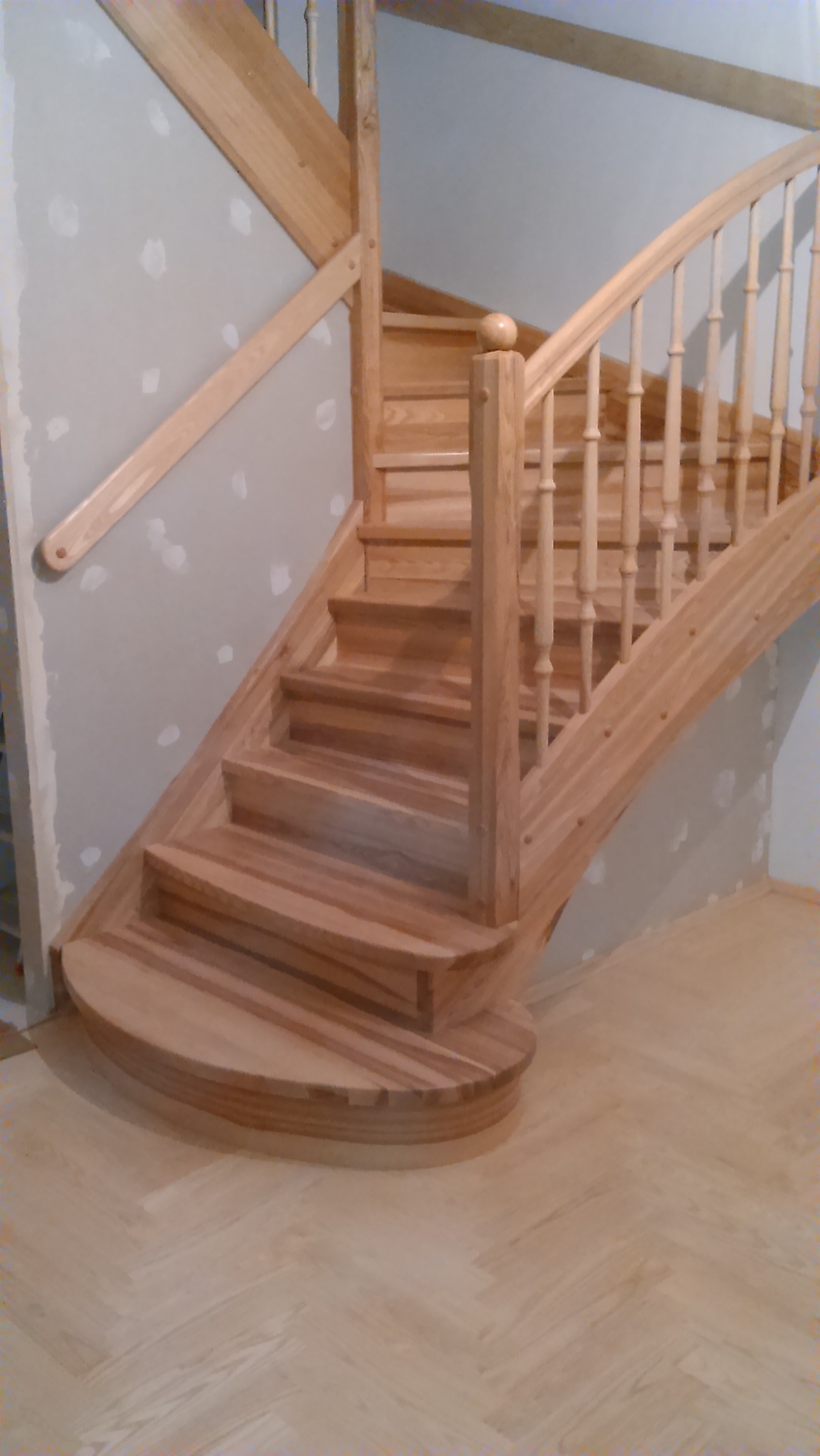 Jasne, drewniane schody z balustradą, widoczne na tle ściany z widocznymi śladami po szpachlowaniu i parkietu ułożonego w jodełkę.