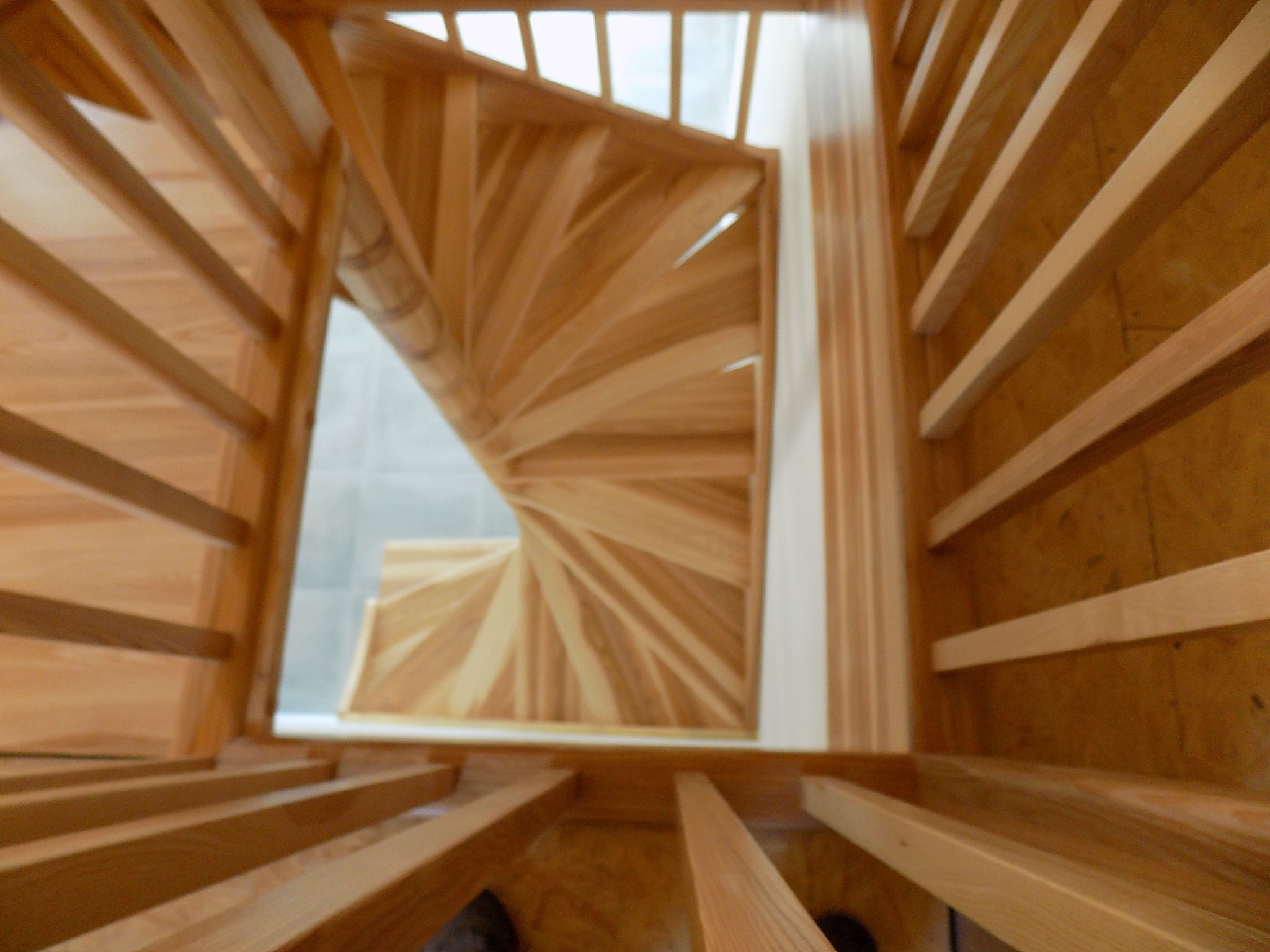 Widok z dołu na spiralne schody z jasnego drewna z balustradą, widoczne fragmenty podłogi na górze, lekko rozmyte.