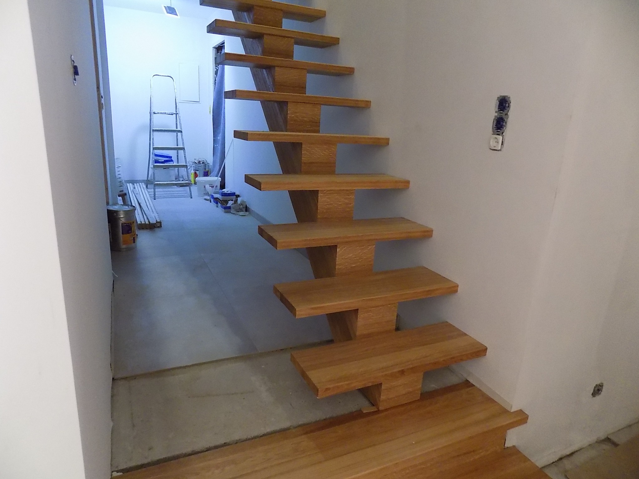 Drewniane schody wspornikowe o jasnym odcieniu drewna, zamontowane przy białej ścianie w trakcie remontu, widoczne puszki elektryczne i drabina w tle.