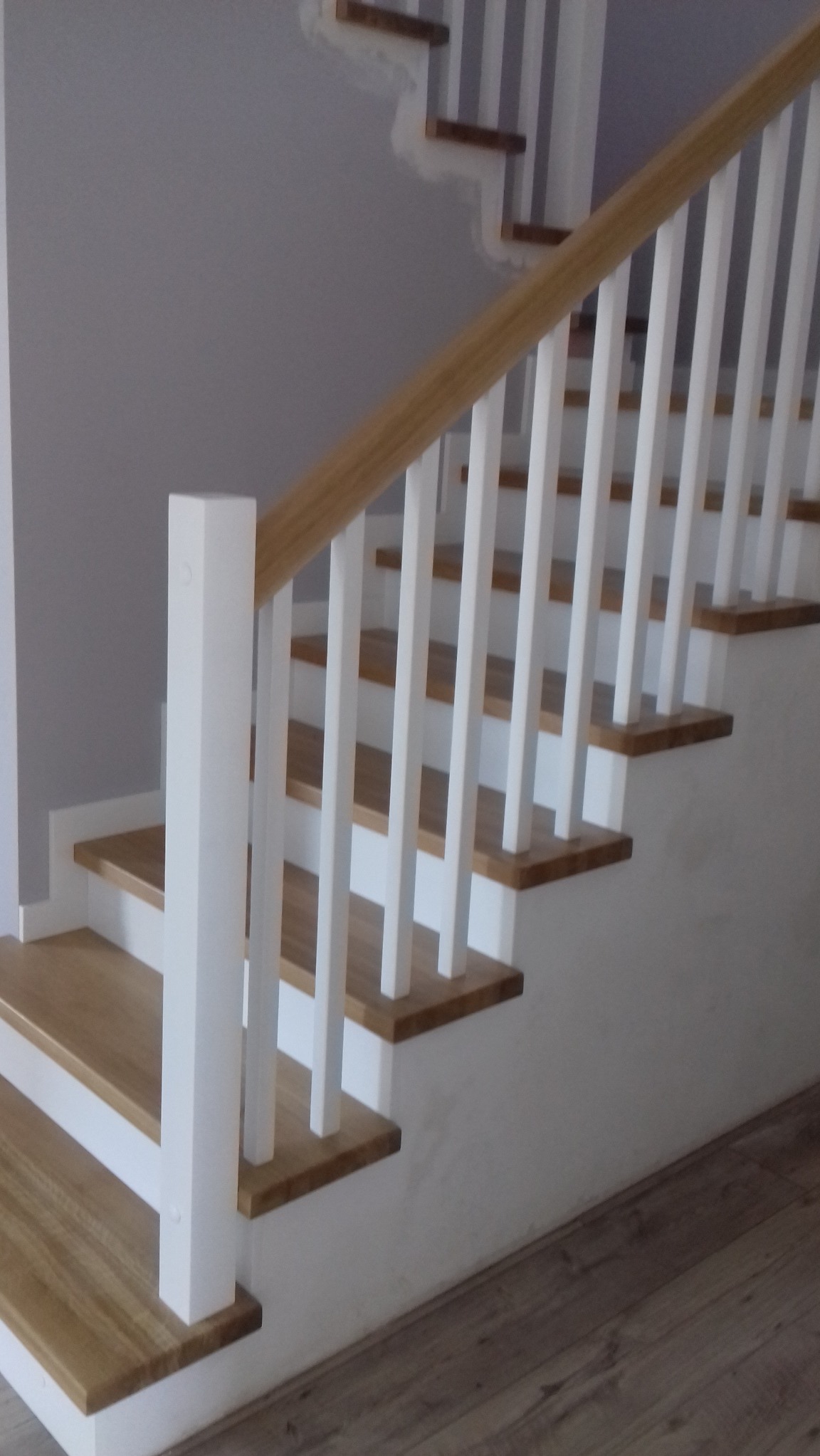 Jasne drewniane stopnie z białymi podstopnicami i balustradą, widok z dołu na konstrukcję przy ścianie pomalowanej na szaro.