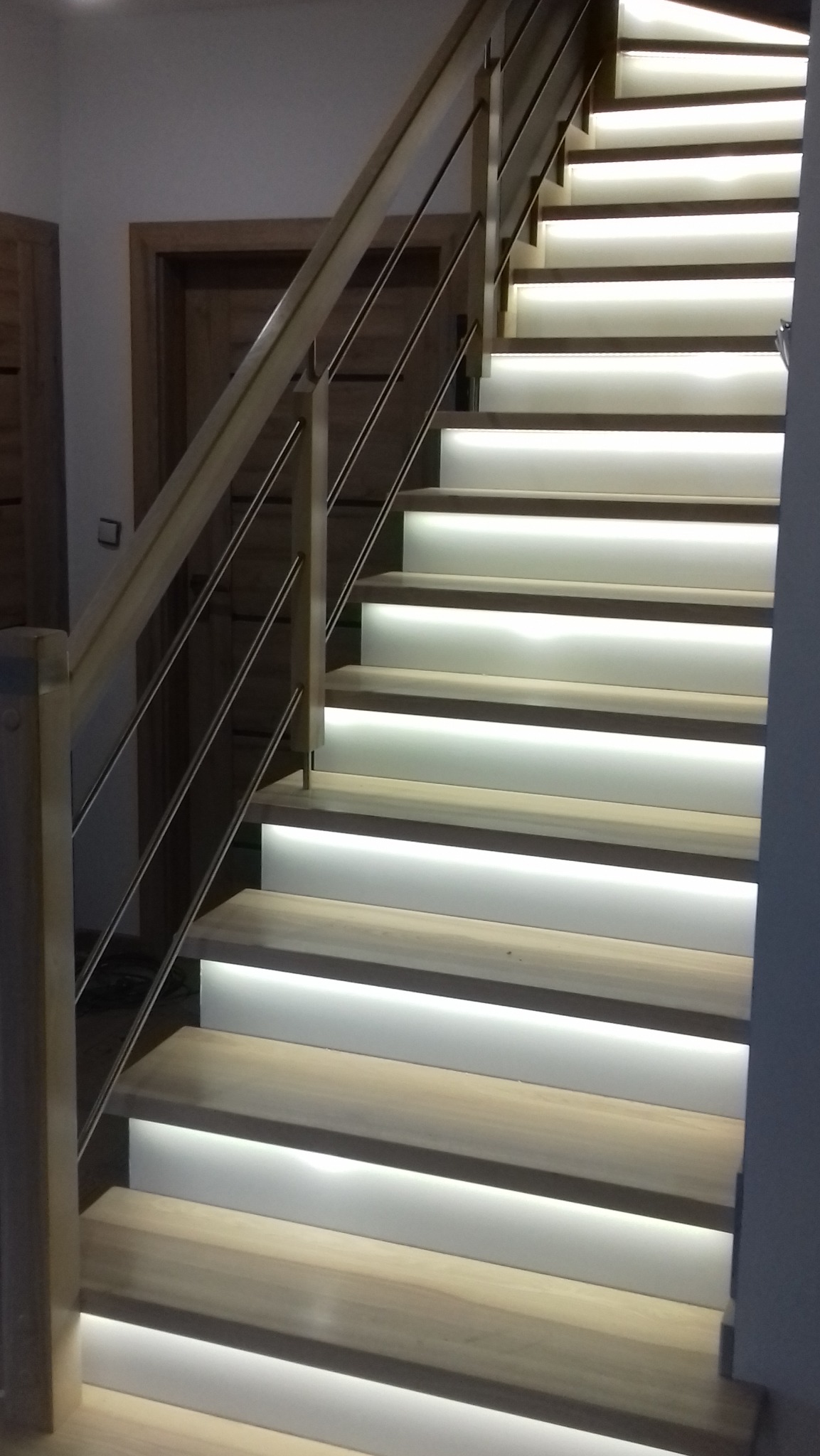 Nowoczesne schody z drewna z podświetleniem LED pod każdym stopniem i metalową balustradą, widok z dołu.