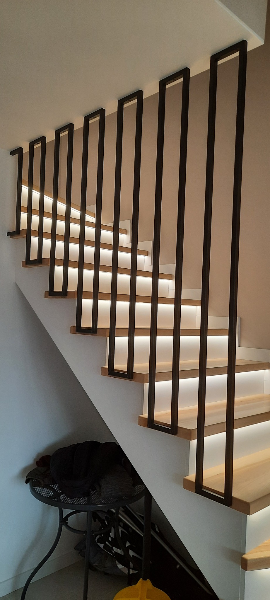 Nowoczesne schody z drewnianymi stopniami, podświetleniem LED i czarną, minimalistyczną balustradą. W tle widoczny stolik z ubraniami.