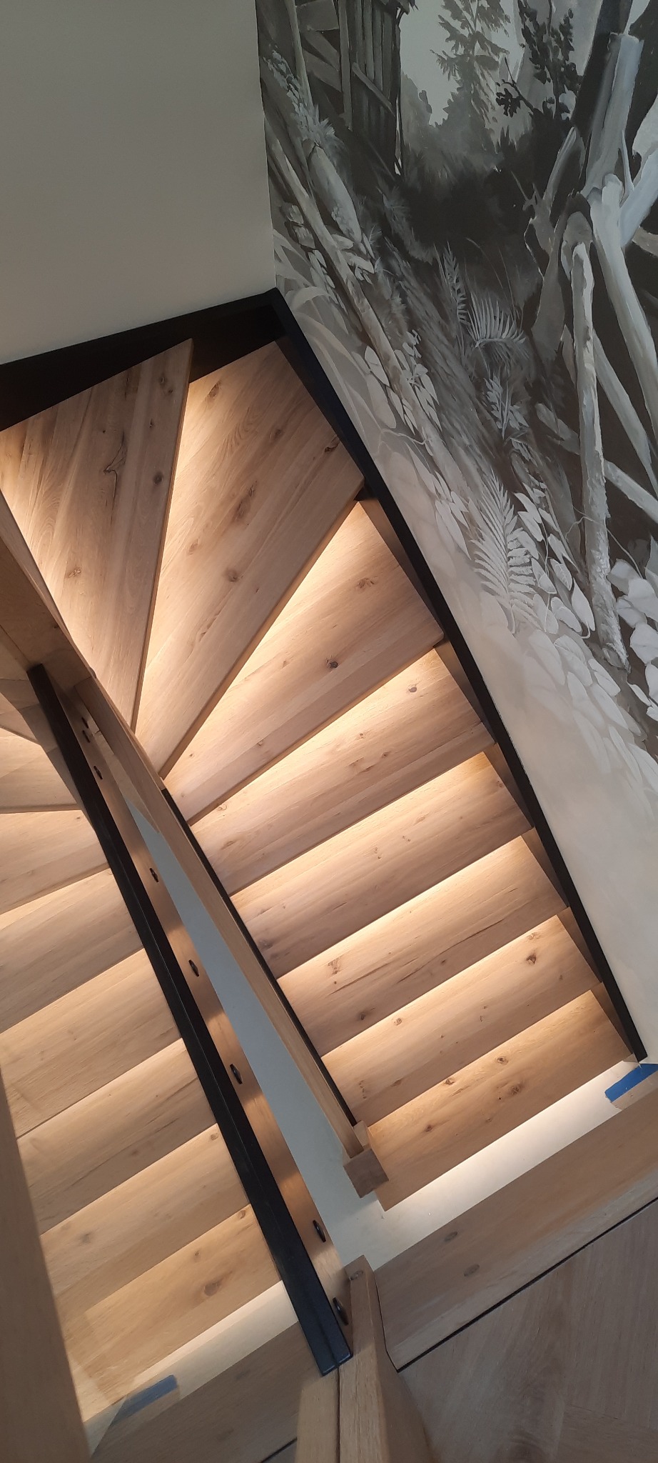 Kątowe ujęcie schodów z jasnego drewna z podświetleniem LED, czarne metalowe elementy konstrukcyjne, ściana z motywem leśnym w odcieniach szarości.