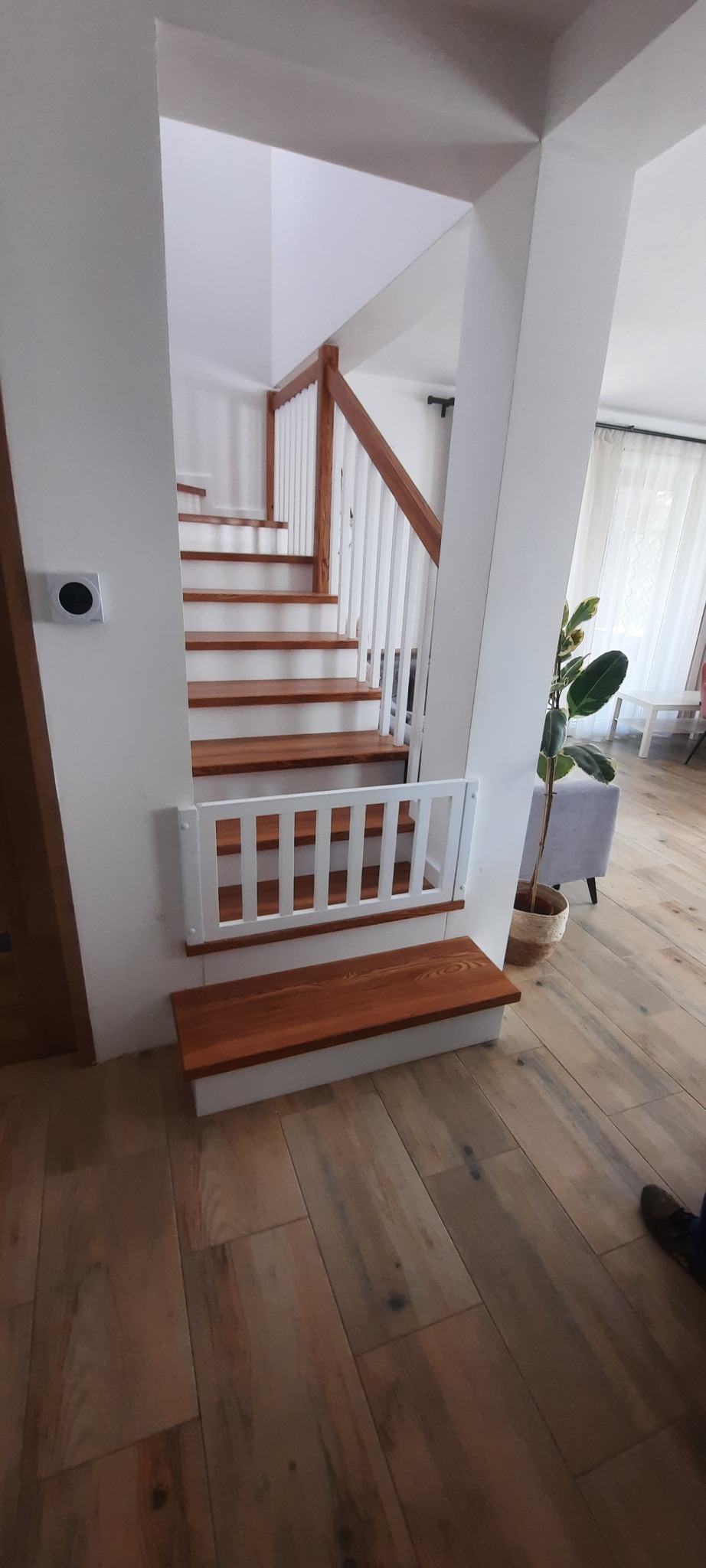 Jasne schody z drewnianymi stopniami i białą barierką, zabezpieczone białą bramką dla dziecka na dole, widok z dołu na drewnianą podłogę.