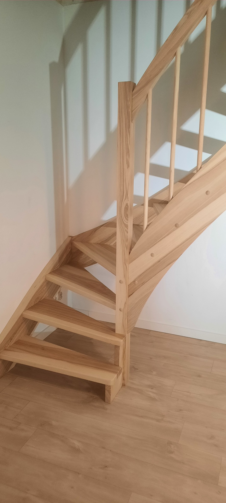 Jasne, drewniane schody z balustradą, zamontowane przy białej ścianie, widoczne od dołu, z parkietem na podłodze.