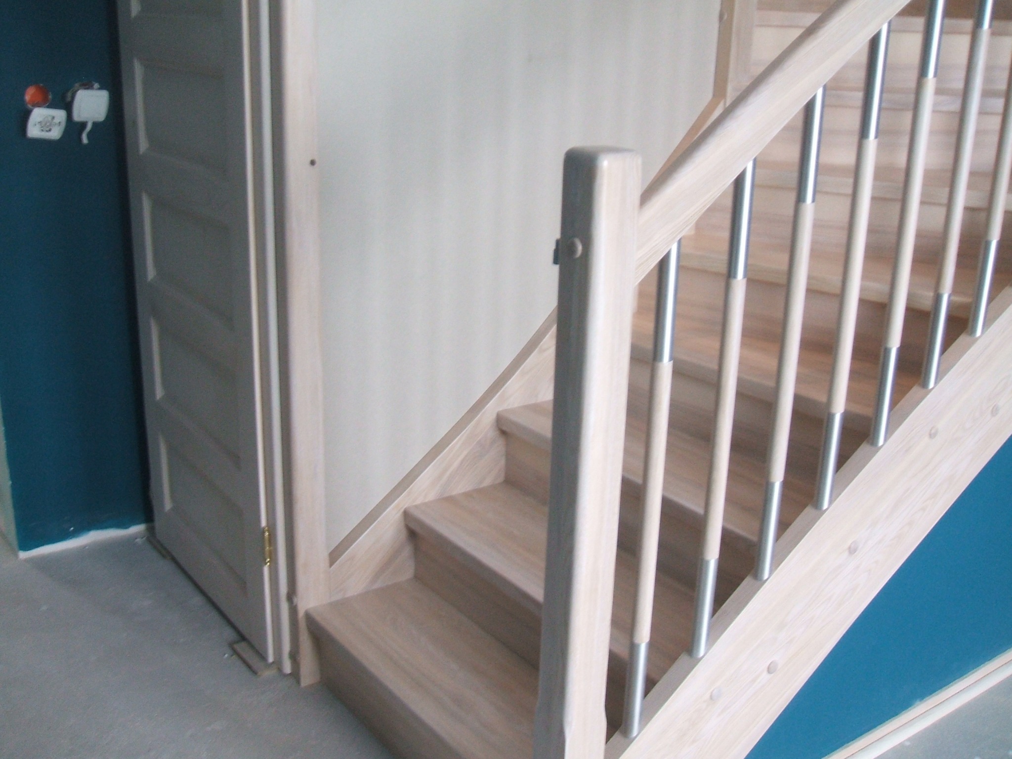 Jasne drewniane schody z metalowymi balustradami, widok od dołu, drzwi w tle, ściana pomalowana na niebiesko.