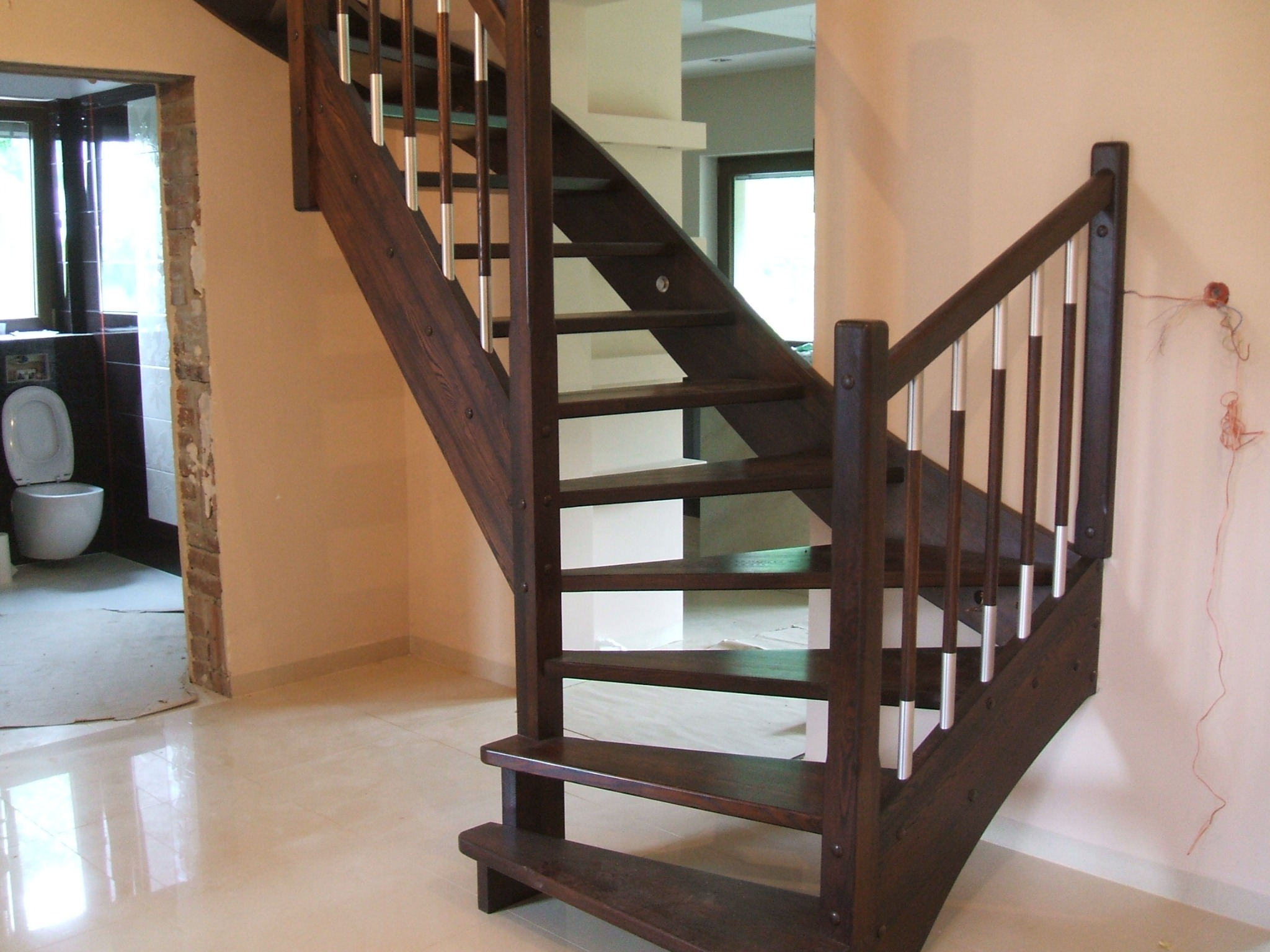Ciemne, drewniane schody z metalowymi balustradami w jasnym wnętrzu, widoczne niedokończone wykończenie ścian i podłogi, oraz fragment łazienki w tle.