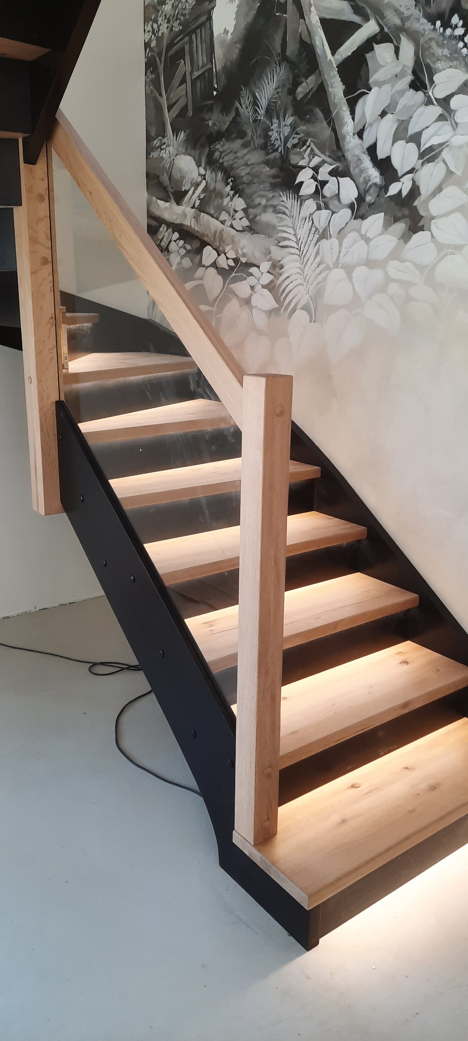 Nowoczesne schody z drewnianymi stopniami podświetlonymi od spodu, czarne metalowe barierki i drewniana poręcz, na ścianie widoczna grafika z motywami roślinnymi.