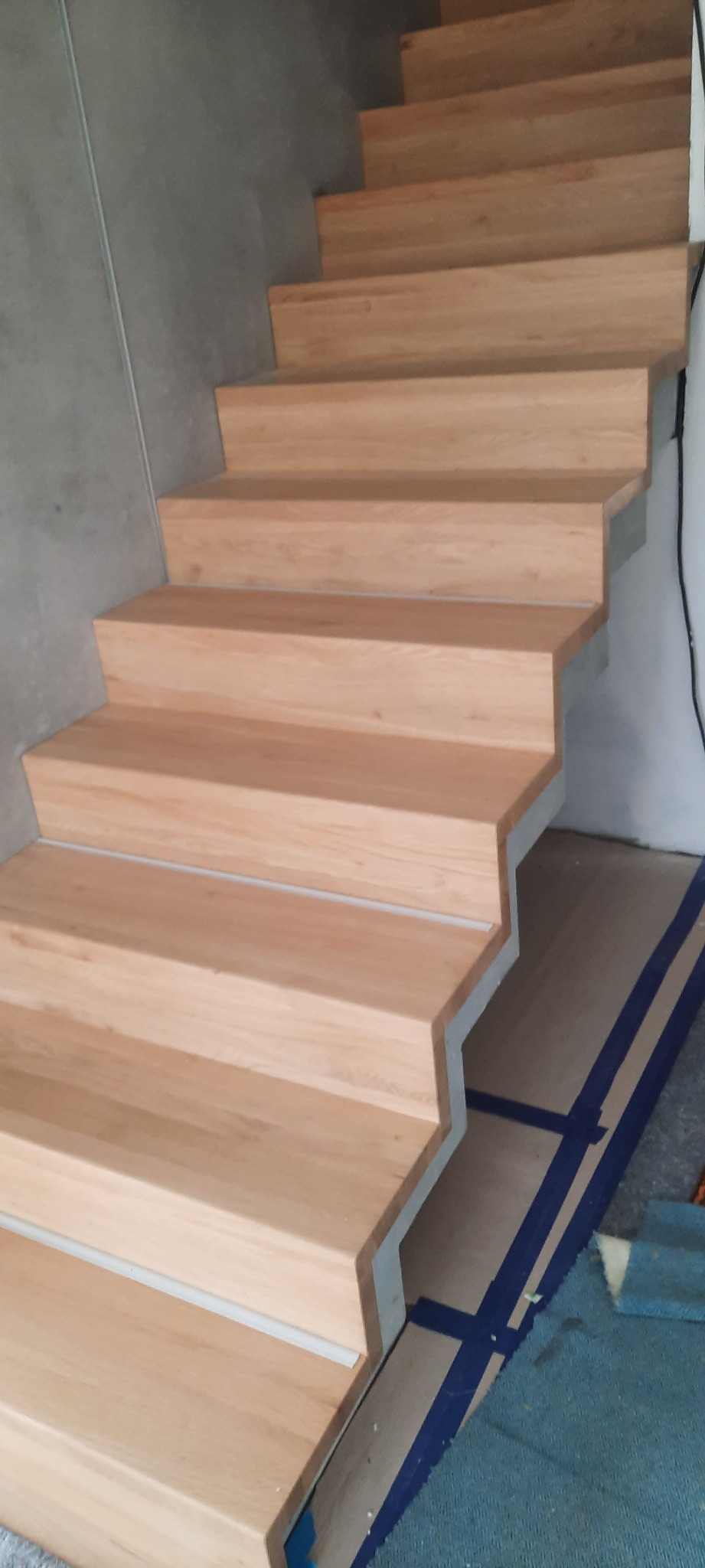 Jasne drewniane schody z widocznym betonowym podkładem i zabezpieczeniem podłogi taśmą malarską, w trakcie montażu.