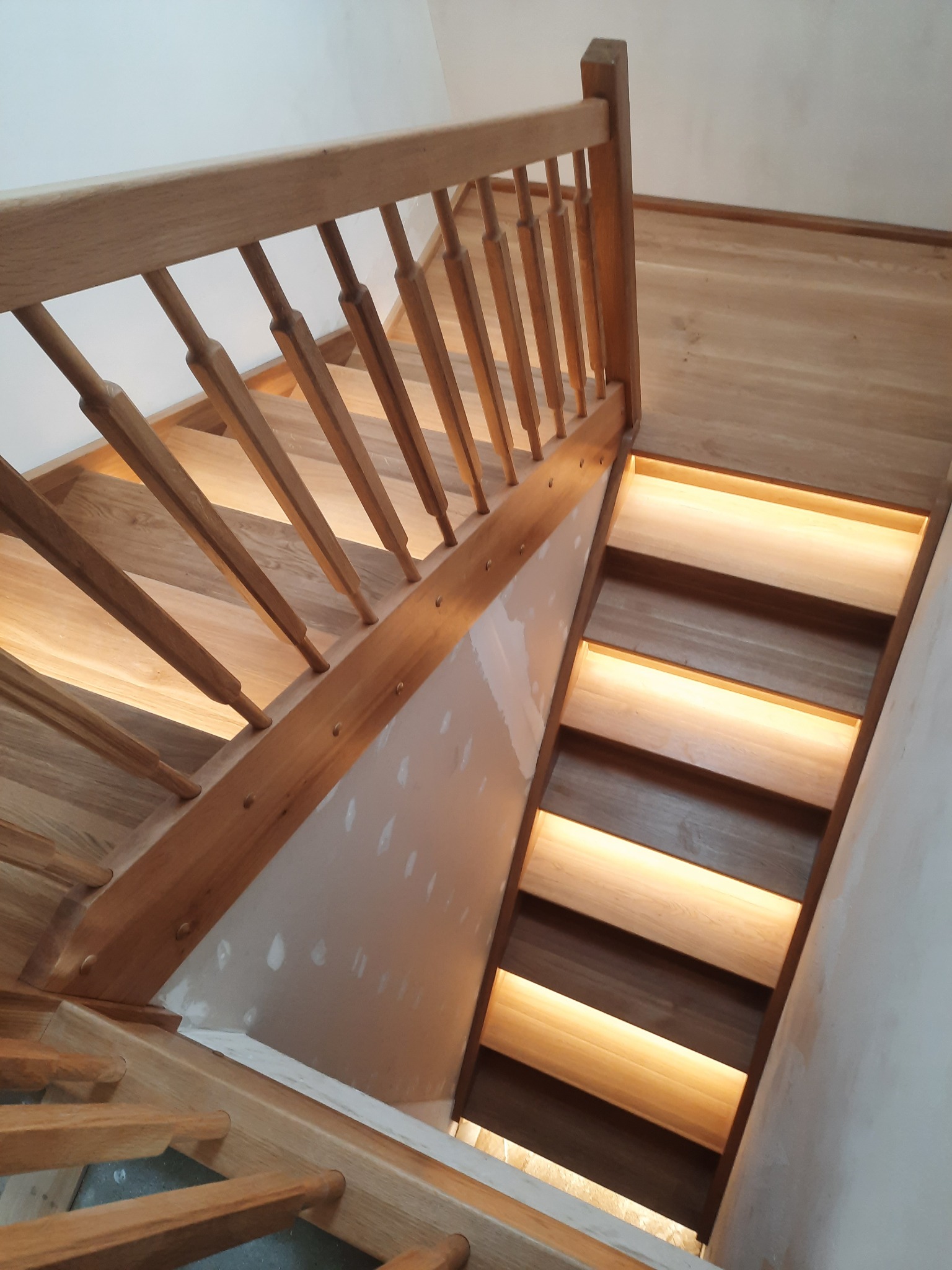 Jasne, drewniane schody z podświetleniem LED na każdym stopniu, widok z góry, ściana w trakcie wykończenia.