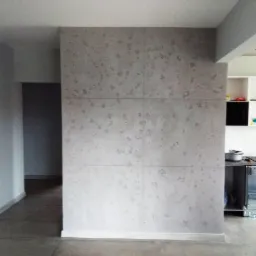 Ściana wykończona betonem architektonicznym w jasnoszarym odcieniu, podzielona na sześć kwadratowych płyt z widocznymi fugami, w tle fragment kuchni i korytarza.